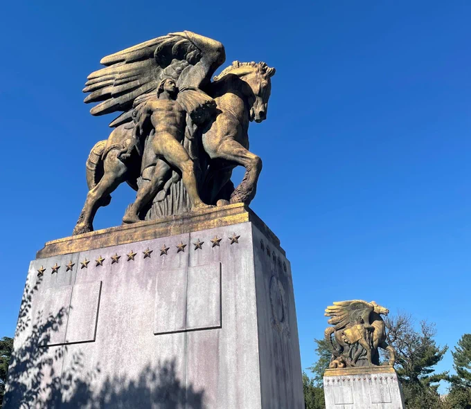 A large bronze sculpture stands atop a tall white stone pedestal against a clear blue sky. The sculpture depicts a winged horse rearing on its hind legs beside a muscular human figure.
