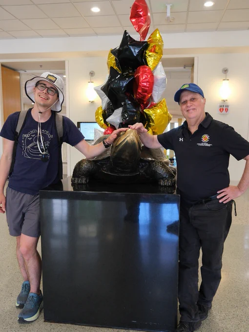 Two people standing on either side of a turtle statue with balloons behind it