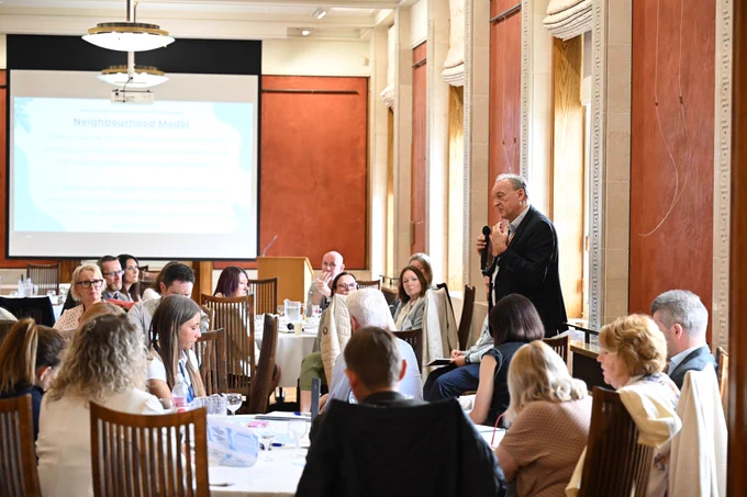 A speaker stands with a microphone addressing attendees seated at round tables in a formal meeting room. A presentation slide titled “Neighbourhood Model” is displayed behind them, while participants listen and review documents at their tables.

