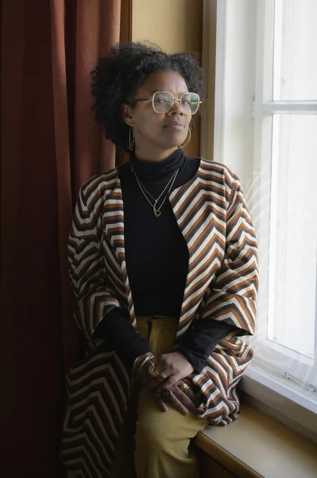 A person wearing glasses and a striped, colorful cardigan sits by a window. They have a thoughtful expression and wear layered necklaces and rings. 

Photo courtesy of Leigh Raiford