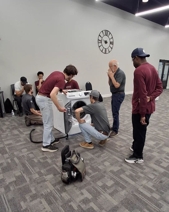 A group of men gathered around a washing machine, inspecting its open door in a spacious room with gray carpet and a wall clock.