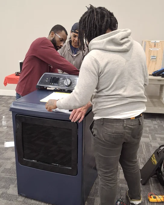 Three individuals examine the control panel of a modern dark blue oven in a room with gray carpet and white walls.