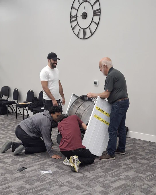 Four men examining a dryer in a carpeted room with chairs and a large wall clock.