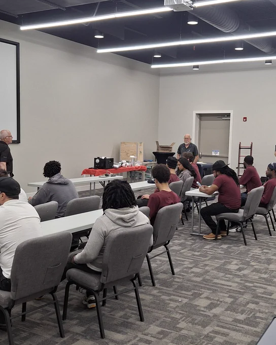 Group of young adults seated in a classroom setting, attentively listening to an instructor standing near a table with equipment and supplies.