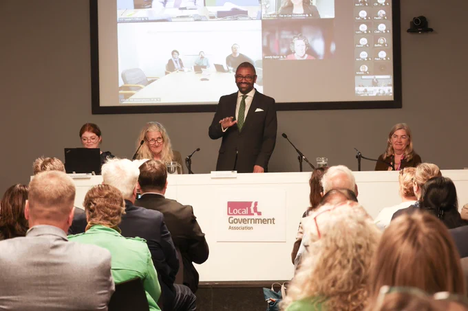Sir James Cleverly speaks at a podium during a Local Government Association event, with a screen showing a video conference behind him.