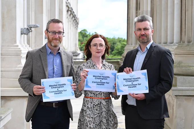 Three people pose outside Parliament Buildings, each holding a sign that reads “Mental Health Champion x Committee for Health.” Stone columns frame the scene, with trees visible behind them.