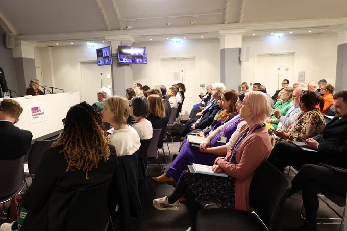 A speaker addresses an audience at a Local Government Association conference, with screens showing remote attendees.
