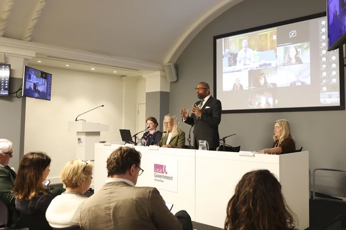 Sir James Cleverly speaks at a podium during a Local Government Association event, with a screen showing a video conference behind him.