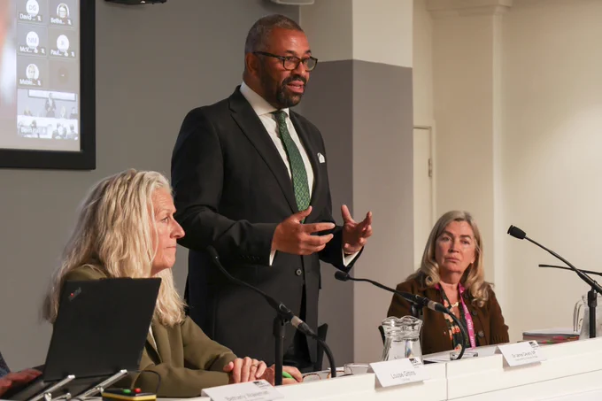 James Cleverly in a suit speaks at a panel discussion with the LGA Chair and CEO seated at the table.