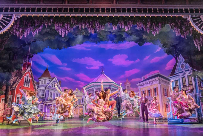A grand theatrical stage production with multiple performers in flowing, colorful costumes dancing across an elaborate set. The backdrop features whimsical Victorian-style buildings\u2014a red church with a steeple on the left, lavender and purple structures in the center and right. Cascading purple wisteria hangs from ornate ceiling details. The sky glows with dramatic blues and pinks, creating a dreamlike, magical atmosphere. Performers in various poses are visible throughout the stage, illuminated by vibrant theatrical lighting.