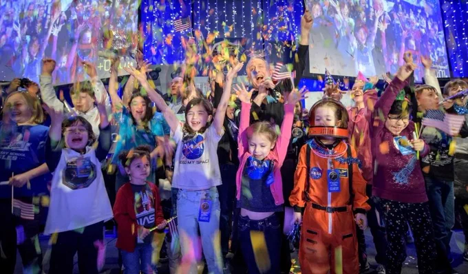 A group of children dressed in NASA gear smiling and holding small American flags with their arms stretched overhead in joy. Colorful confetti falls from above and the room is dark blue.