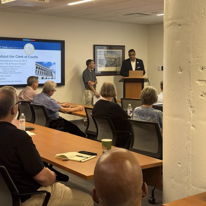 A man in a dark blazer stands at a wooden podium presenting to a small audience in a conference room. To his left, another man stands near a large wall-mounted display screen showing a slide titled "About the Clerk of Courts" with a list of divisions including Administration–Finance, HR, IT; Auto Title & Passport Division; Common Pleas Division; Court of Appeals; Municipal Court Division; Help Center; Bailiffs; and Central Services Division.  Several attendees — men and women of varying ages — are seated around a long wooden conference table with notebooks, water bottles, and coffee cups.