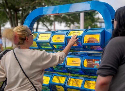 A woman in a light sweater interacts with a blue, interactive, word-display installation outdoors. Her expression is thoughtful and engaged.
