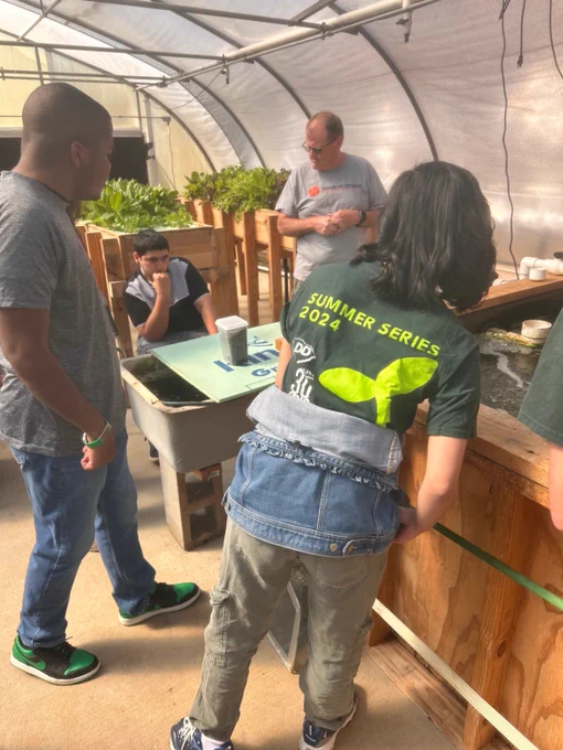 A group of people stand in a greenhouse around a water tank. One person wears a green shirt with "SUMMER SERIES 2024" and a seedling logo on the back.