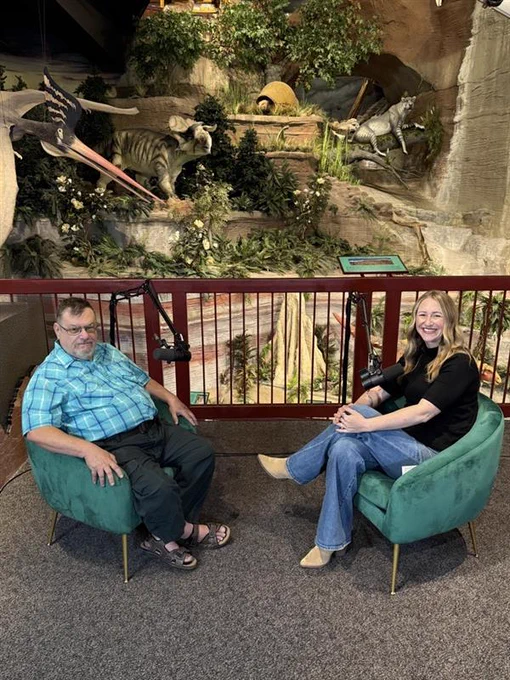 Dr. Bob and Ashley are seated in green chairs facing each other with a detailed prehistoric diorama featuring a saber-toothed tiger and other ancient animals in the background.