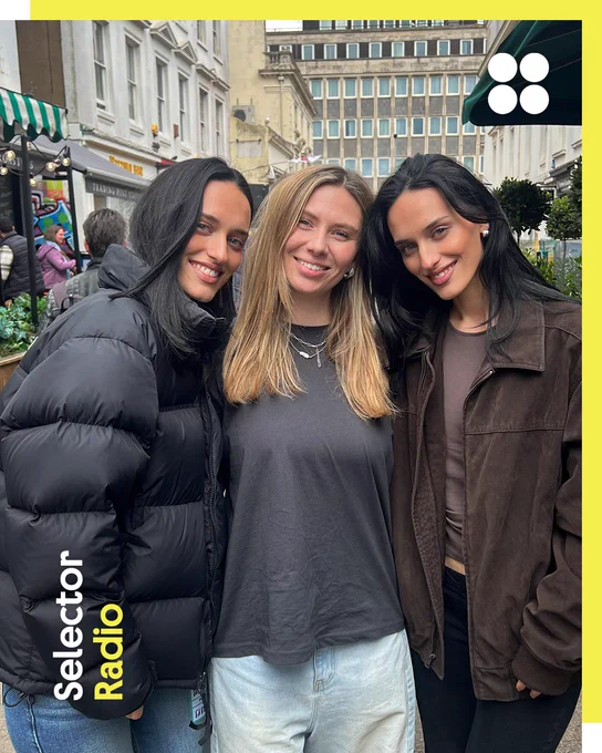 Three women, the duo Dolder are either side of presenter Sian, are smiling and posing together outdoors. Dolder, Dani and Zara are identitical twins. They are standing in a street with buildings and greenery in the background. The leftmost woman is wearing a black puffer jacket, the middle woman is in a grey top, and the rightmost woman is dressed in a brown jacket. The image has a yellow border with the text "Selector Radio" in the bottom left corner.
