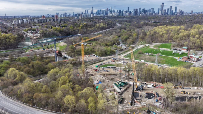 Aerial view of a construction site surrounded by trees, with cranes and equipment visible. The site is set in a wooded area with a sprawling city skyline in the background, including the CN Tower and other Toronto landmarks. Roads and pathways intersect the landscape. 