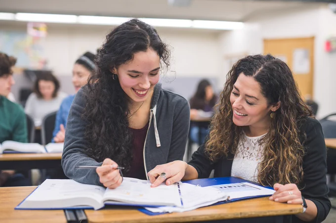 A student and teacher in a classroom are sitting at a desk, smiling and looking at a book together. Other students are visible in the background.