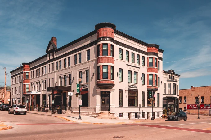A large historic corner building with rounded brick bays and a “1899” date marker stands at a downtown intersection under a blue sky.