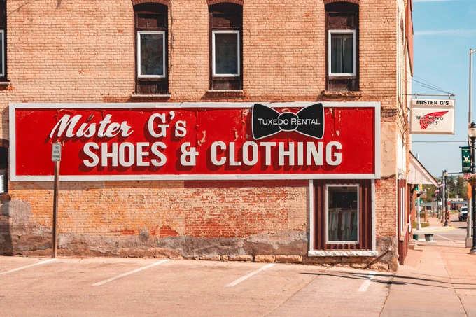 A weathered brick building displays a large red “Mister G’s Shoes & Clothing” sign with a tuxedo rental logo, set beside a small parking area and downtown sidewalk.