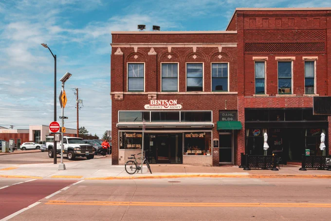 A red brick historic downtown storefront with a “Bentson Insurance Agency” sign faces a wide street, with a bicycle parked near the entrance and neighboring brick buildings extending to the right.