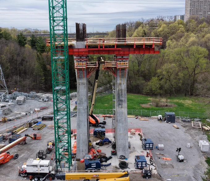 A construction site with a large green crane and two tall concrete columns, supporting a red steel beam structure at the top. Workers and machinery are visible around the site.