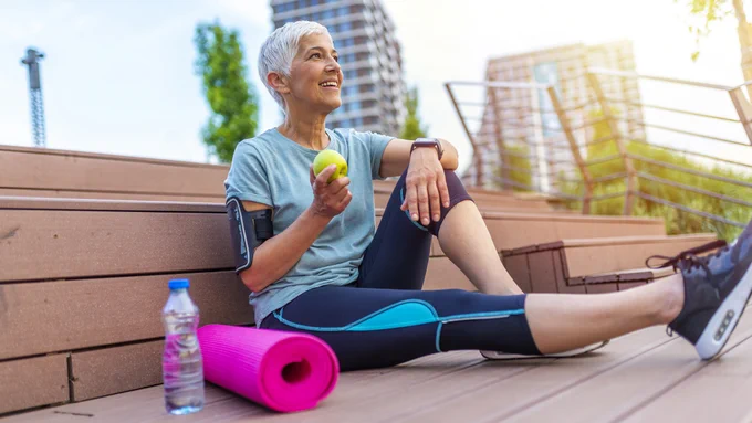 Sport mature woman resting after exercise and eating apple outside. 