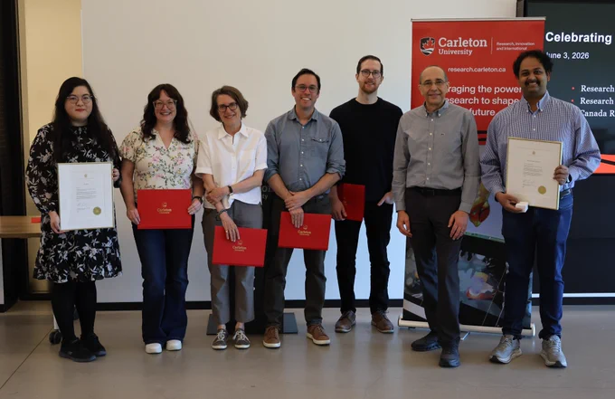 Seven people posing for a photo. Four are holding red folders, and two are holding framed letters. Carleton-branded banner in the background.