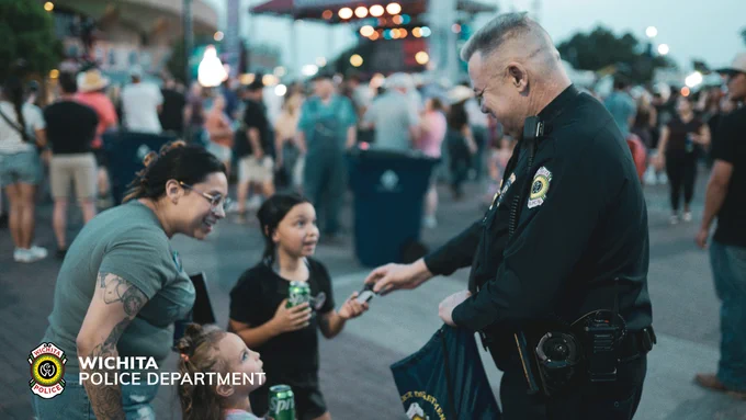 A law enforcement officer from the Wichita Police Department interacts with two children at an outdoor event. One child is holding a canned drink. There is a crowd in the background and a stage with lights. The department's logo is visible in the bottom left corner.