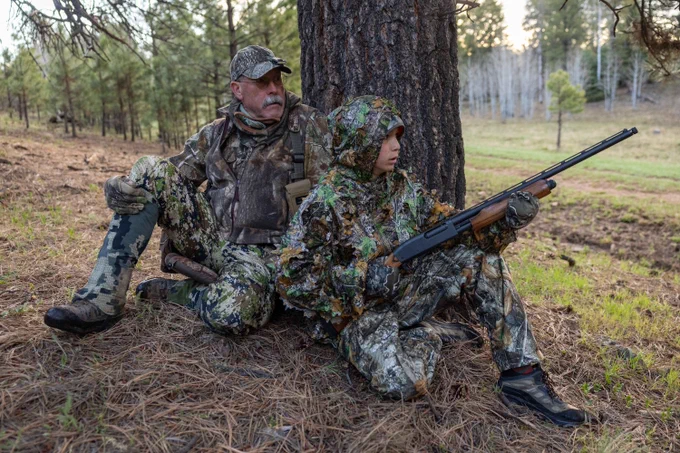 A man and teenager dressed in camouflage clothes sit beneath a large pine tree in the morning, the teenager is holding a shotgun.