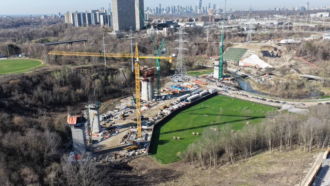  Un chantier de construction en cours avec plusieurs grues et des structures en béton. Le chantier est entouré d’arbres et de champs ouverts. Une rivière à proximité coule le long du site. À l’arrière-plan, il y a de nombreux immeubles de grande hauteur et une ligne d’horizon de la ville avec la Tour CN visible.