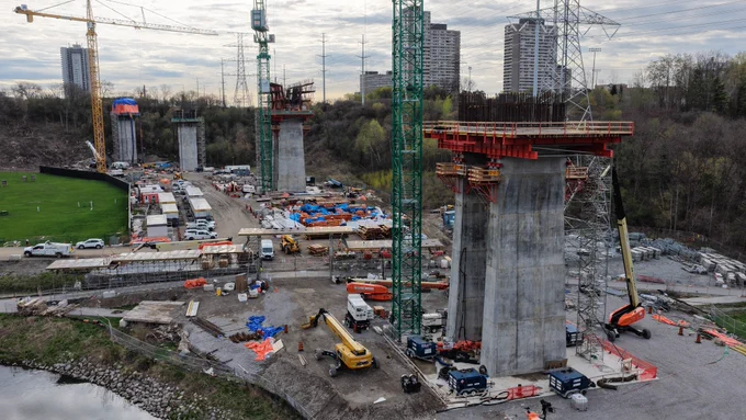 Un chantier de construction en cours avec plusieurs grands piliers en béton et entouré de plusieurs grues et véhicules de construction. La zone est vaste, avec divers matériaux éparpillés, et en arrière-plan se dressent des immeubles de grande hauteur sur fond de ciel nuageux.