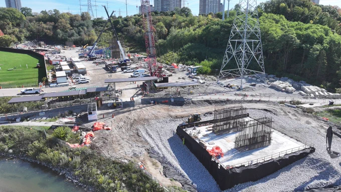 Vue aérienne d’un chantier de construction avec des grues, des structures en acier et des machines. Un grand pylône d’électricité se dresse de manière proéminente. L’arrière-plan comprend des arbres, un terrain de sport et plusieurs bâtiments, y compris quelques grandes structures. Le gravier et les matériaux de construction sont répartis dans toute la zone.