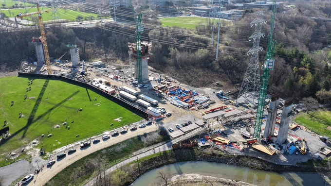 Un chantier de construction avec plusieurs grandes grues et des structures partielles de pont est visible. La zone est entourée de champs verts et d’un cours d’eau. À proximité, il y a des travailleurs et des véhicules stationnés.