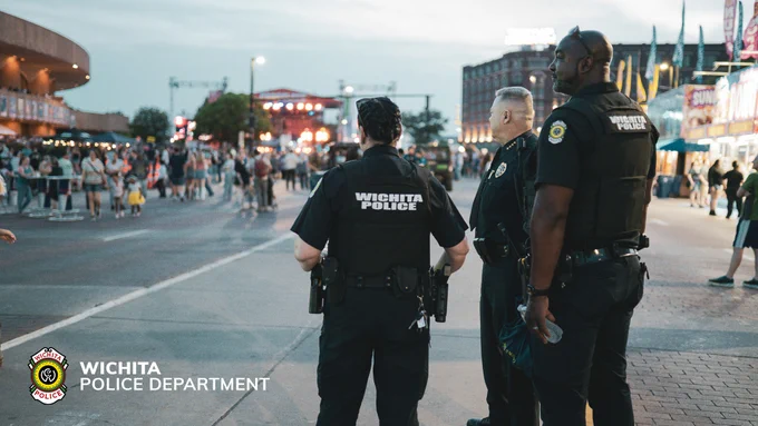 Police officers from the Wichita Police Department stand together in a busy street setting with people walking in the background. The department logo is visible in the bottom left corner.