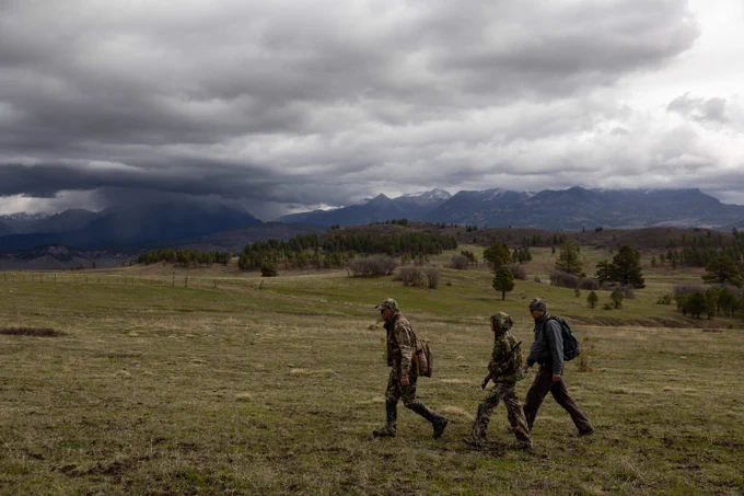 Three people dressed in camouflage clothes, one of them carrying a shotgun, walk across a large grassy field with storm clouds and large mountains in the background.