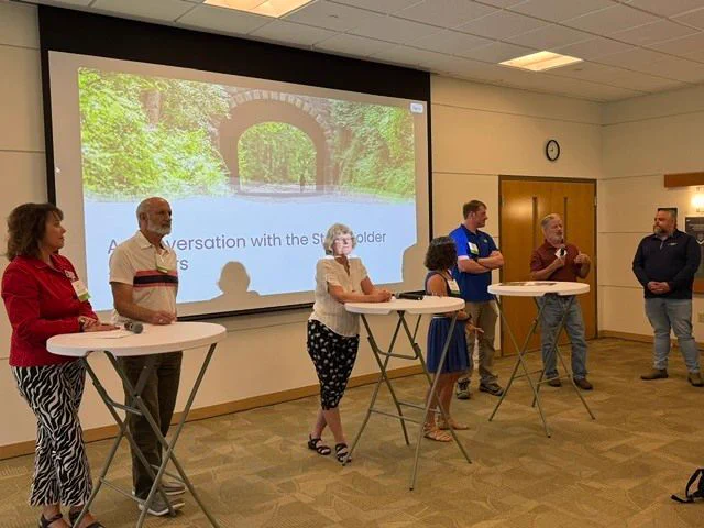 Seven people presenting at the front of a conference room. There is a powerpoint playing in the background. 