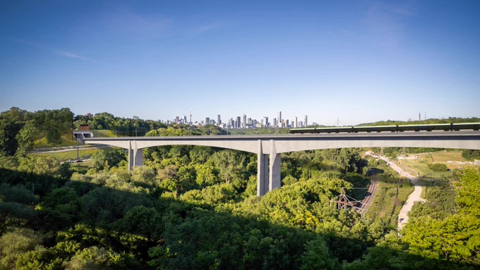 Aerial view of a cityscape featuring a large highway viaduct stretching across a lush, green landscape with autumn foliage. High-rise buildings are visible in the background under a clear blue sky.