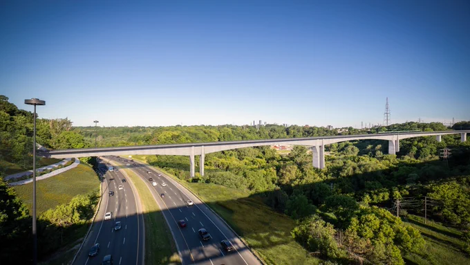 Vue aérienne d’une autoroute avec plusieurs voies de circulation, entourée d’arbres verts luxuriants et d’herbes. Un long pont surélevé traverse l’autoroute. Le ciel est clair et bleu.