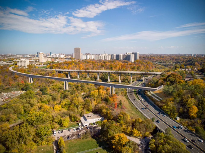  Vue aérienne d’un paysage urbain présentant un grand viaduc d’autoroute s’étendant sur un paysage luxuriant et vert avec des feuillages d’automne. Des immeubles de grande hauteur sont visibles en arrière-plan sous un ciel bleu clair.
