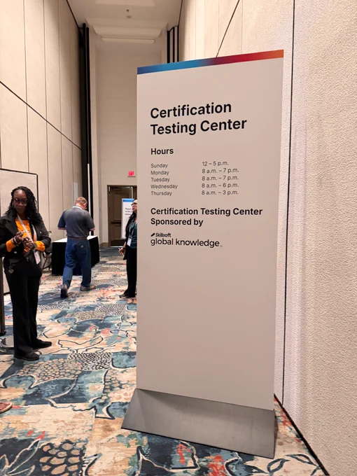 A white sign for a Certification Testing Center, listing daily hours from Sunday (12-5 pm) to Thursday (8 am - 3 pm). It's sponsored by Skillsoft global knowledge. People are visible in the background, including a woman checking her phone on patterned carpet.