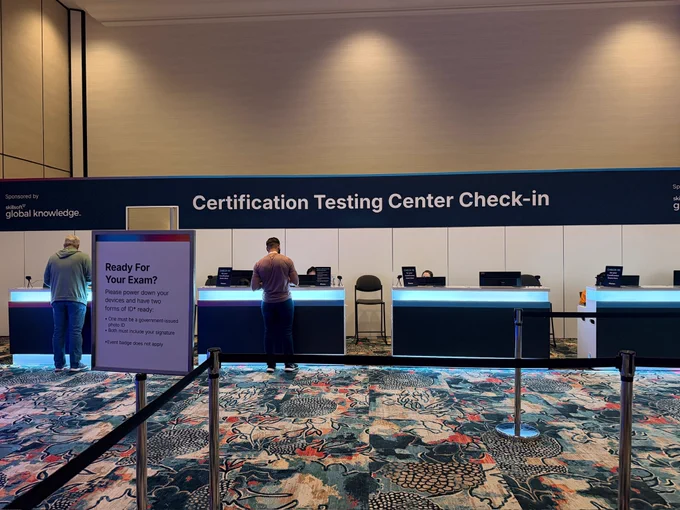 A Certification Testing Center Check-in area with a large banner displaying 'Certification Testing Center Check-in'. Two people are at separate counters. A white sign in the foreground reads, 'Ready For Your Exam? Please power down your devices and have two forms of ID* ready: government-issued photo ID & signature needed.' The area is sponsored by Skillsoft.