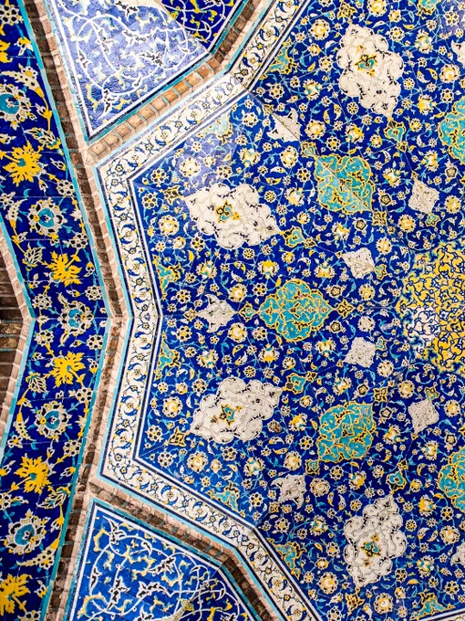 An overhead, full-frame shot of an intricate Safavid-era mosaic tile ceiling from the Sheikh Lotfollah Mosque in Isfahan, Iran. The symmetrical, radial pattern expands outward from a central golden-yellow medallion. The design features a complex web of swirling floral arabesques and botanical motifs that unfold in layers of cobalt blue, turquoise, and gold, where geometry and floral motifs converge into a mesmerizing expression of symmetry, light, and devotion.