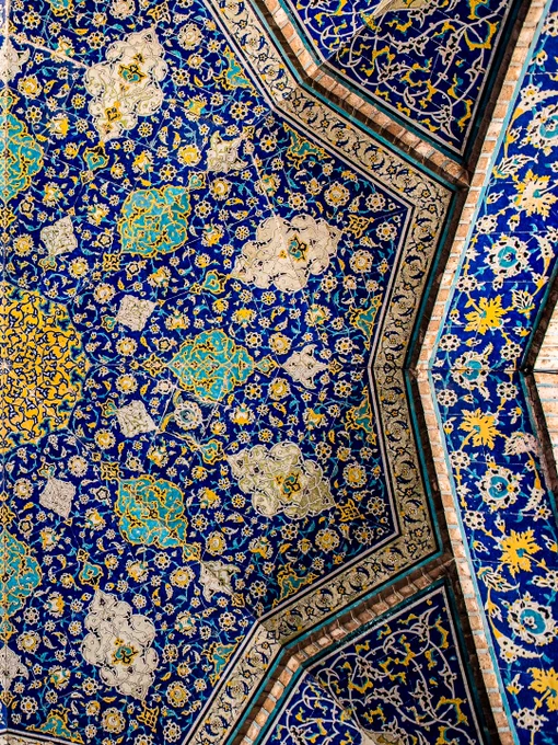An overhead, full-frame shot of an intricate Safavid-era mosaic tile ceiling from the Sheikh Lotfollah Mosque in Isfahan, Iran. The symmetrical, radial pattern expands outward from a central golden-yellow medallion. The design features a complex web of swirling floral arabesques and botanical motifs that unfold in layers of cobalt blue, turquoise, and gold, where geometry and floral motifs converge into a mesmerizing expression of symmetry, light, and devotion.