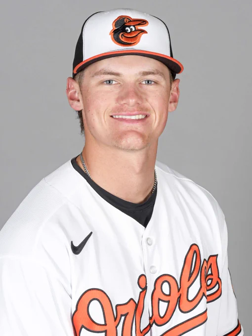 Nate George looks at the camera and smiles. He is wearing a white Orioles jersey and a tri-color cap.