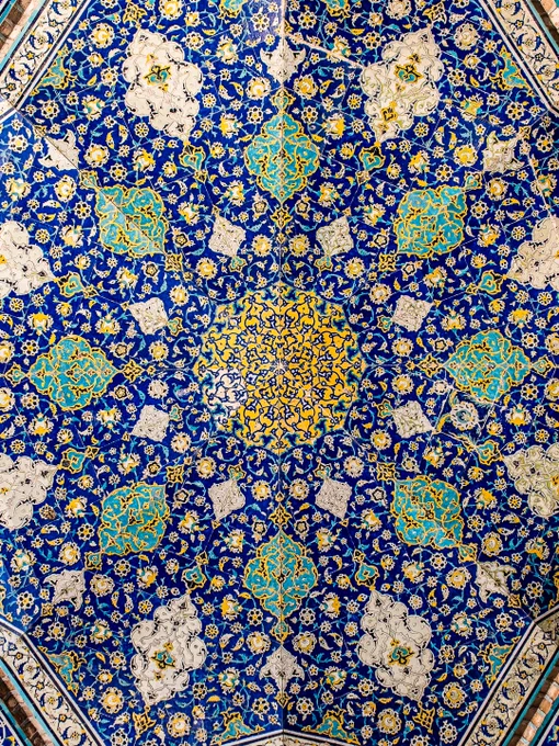 An overhead, full-frame shot of an intricate Safavid-era mosaic tile ceiling from the Sheikh Lotfollah Mosque in Isfahan, Iran. The symmetrical, radial pattern expands outward from a central golden-yellow medallion. The design features a complex web of swirling floral arabesques and botanical motifs that unfold in layers of cobalt blue, turquoise, and gold, where geometry and floral motifs converge into a mesmerizing expression of symmetry, light, and devotion.