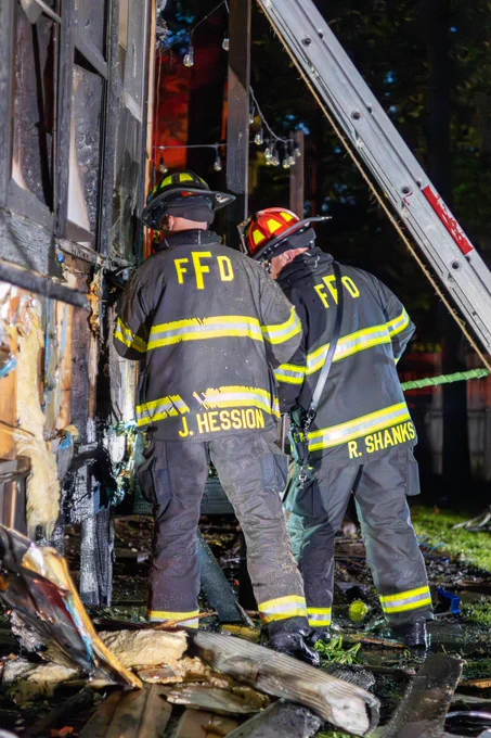 Two Fishers firefighters work beside a fire-damaged exterior wall and sunroom area of a home. A ground ladder is positioned against the structure as crews check for remaining hot spots and fire extension.