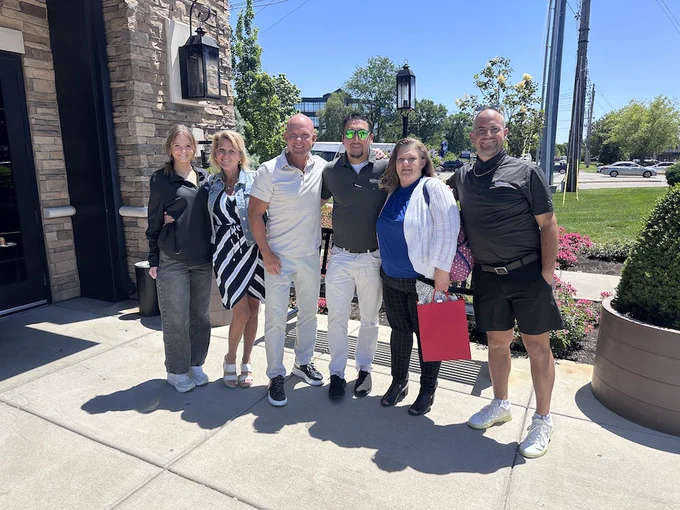 Group of six adults standing together outside a stone building on a sunny day, dressed in casual and semi-formal attire.