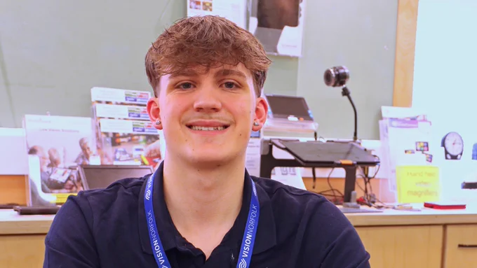 Volunteer Max sitting in the Norwich Equipment Centre with pamphlets and low vision equipment behind him.