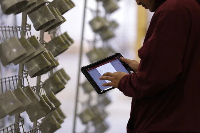 A person in a maroon shirt uses a tablet, standing near a rack of gray powder-coated items, likely in a manufacturing setting.